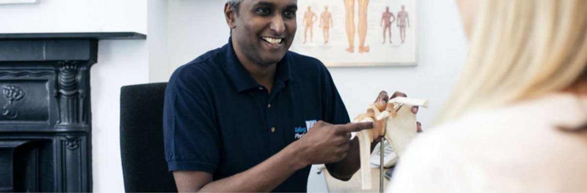 Wide shot of Karthik holding a type of human bone model whilst smiling at a patient.